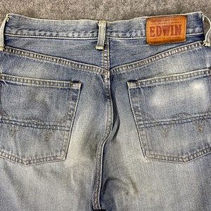 May include: Close-up of the back of blue denim jeans. The jeans have two back pockets and a brown leather patch with the brand name "EDWIN". The denim has a faded, worn look.