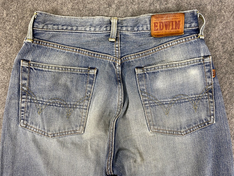 May include: Close-up of the back of blue denim jeans. The jeans have two back pockets and a brown leather patch with the brand name "EDWIN". The denim has a faded, worn look.