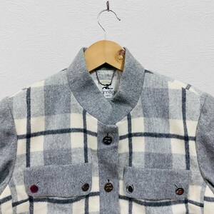 K&ouml;nnte beinhalten: Grau-wei&szlig;-schwarz karierte Jacke mit grauem Kragen und Taschen. Die Jacke hat drei dekorative Kn&ouml;pfe auf der Vorderseite und zwei auf den Taschen. Der Markenname "urr&acirc;ge paris" ist auf einem Etikett in der N&auml;he des Kragens zu sehen.