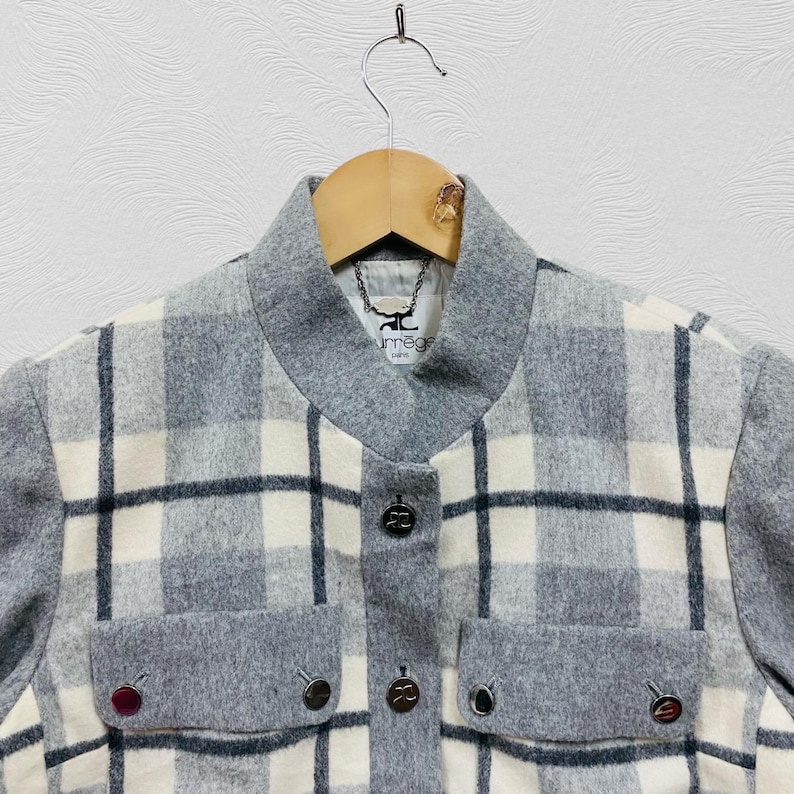 K&ouml;nnte beinhalten: Grau-wei&szlig;-schwarz karierte Jacke mit grauem Kragen und Taschen. Die Jacke hat drei dekorative Kn&ouml;pfe auf der Vorderseite und zwei auf den Taschen. Der Markenname "urr&acirc;ge paris" ist auf einem Etikett in der N&auml;he des Kragens zu sehen.