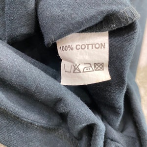 Pode incluir: Close-up de um tecido azul escuro com uma etiqueta branca. A etiqueta diz "100% COTTON" em texto preto. Abaixo do texto est&atilde;o v&aacute;rios s&iacute;mbolos de lavagem.