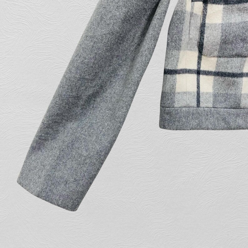 K&ouml;nnte beinhalten: Graue Wolljacke mit Karomuster auf der Vorderseite und einem einfarbigen grauen &Auml;rmel. Das Karomuster zeigt wei&szlig;e und marineblaue Quadrate. Die Jacke hat einen klassischen, ma&szlig;geschneiderten Look.