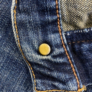 May include: Close-up of a blue denim fabric with a gold button and brown stitching.