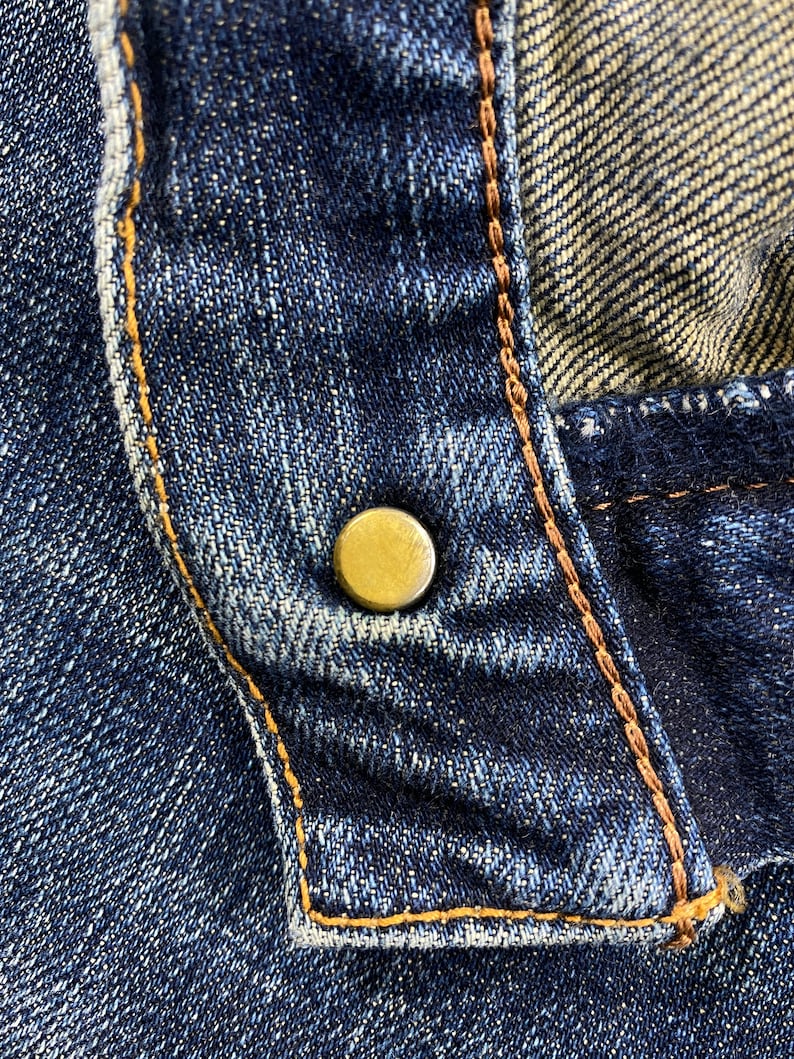 May include: Close-up of a blue denim fabric with a gold button and brown stitching.