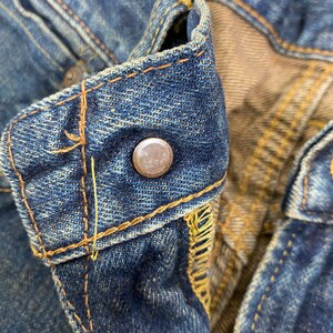 May include: Close-up of a pair of blue denim jeans, focusing on the button closure and stitching. The jeans show signs of wear and fading, with visible yellow stitching and a metal button. A small orange tag is visible.