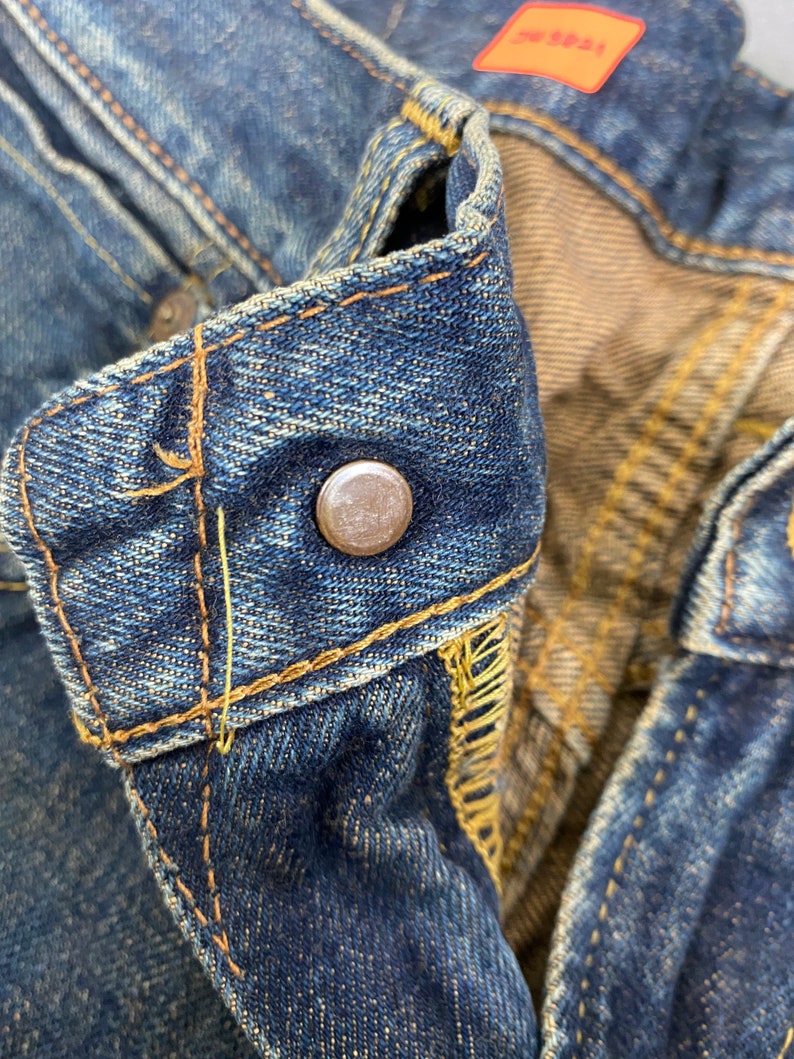May include: Close-up of a pair of blue denim jeans, focusing on the button closure and stitching. The jeans show signs of wear and fading, with visible yellow stitching and a metal button. A small orange tag is visible.