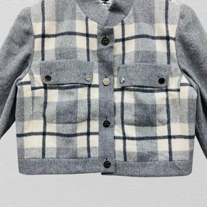 K&ouml;nnte beinhalten: Grau-wei&szlig; karierte Jacke mit rundem Ausschnitt und langen &Auml;rmeln. Die Jacke hat zwei Vordertaschen und dekorative Kn&ouml;pfe vorne. Das Karomuster ist eine Mischung aus grauen, wei&szlig;en und schwarzen Linien.