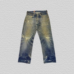 Könnte beinhalten: Blaue Jeans aus Denim mit abgenutzter Optik, sichtbaren Verwaschungen, Sitzfalten an den Hüften und ausgefransten Rissen an den Oberschenkeln. Die Hose verfügt über einen Knopfverschluss, fünf Taschen und ein kleines Loch am linken Beinabschluss.