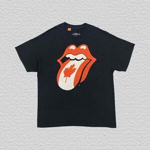 May include: Black t-shirt with the Rolling Stones' tongue logo, incorporating a Canadian maple leaf. The tongue is red and white, with the maple leaf at the centre. A classic rock and roll tee.