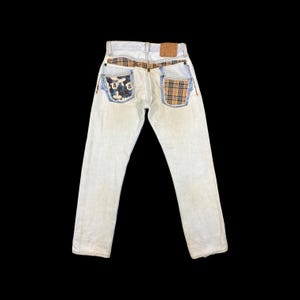 May include: Light wash denim jeans with unique patchwork details. The back pockets feature contrasting patterns, including a Mickey Mouse print and a plaid design. A leather patch is visible at the waistband.