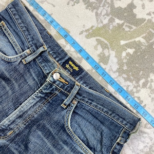May include: Blue denim jeans with a brown leather patch that says "Wrangler" on the back waistband. The jeans have a light blue wash and a faded look.