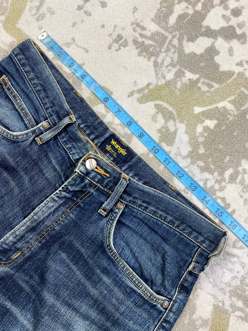 May include: Blue denim jeans with a brown leather patch that says "Wrangler" on the back waistband. The jeans have a light blue wash and a faded look.