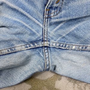 K&ouml;nnte beinhalten: Nahaufnahme einer hellblauen Jeans. Das Bild konzentriert sich auf die Vorderseite der Jeans und zeigt den Knopfverschluss, die N&auml;hte und die Textur des Stoffes. Der Denim hat ein abgenutztes Aussehen.