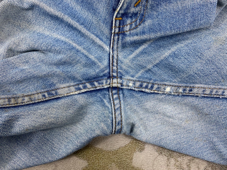 K&ouml;nnte beinhalten: Nahaufnahme einer hellblauen Jeans. Das Bild konzentriert sich auf die Vorderseite der Jeans und zeigt den Knopfverschluss, die N&auml;hte und die Textur des Stoffes. Der Denim hat ein abgenutztes Aussehen.