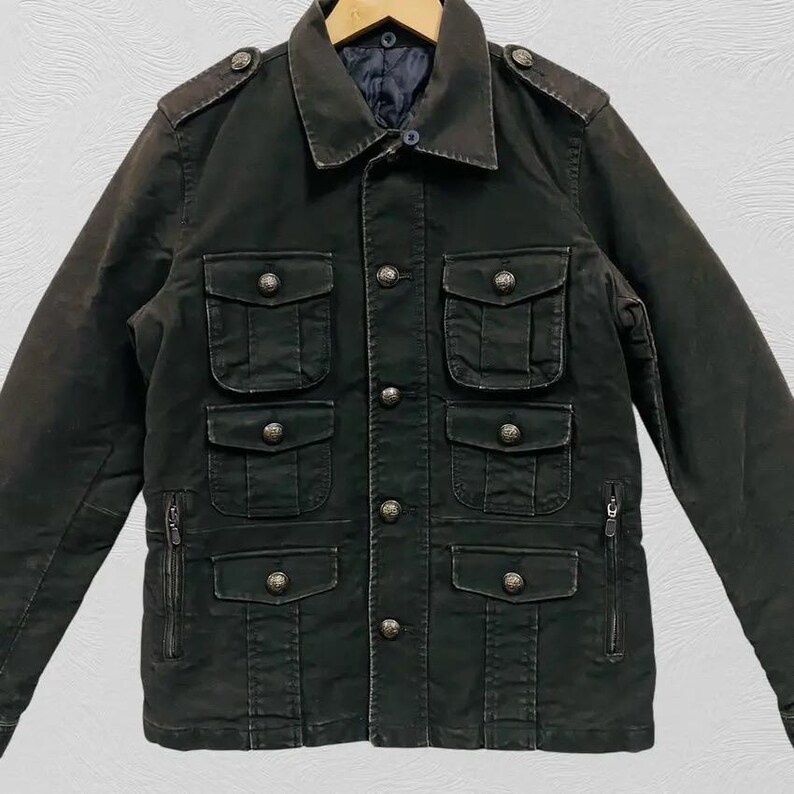 May include: Dark olive green jacket with multiple front pockets and decorative silver buttons. The jacket has a collared neckline, epaulets, and zippered side pockets. The jacket is made of a durable fabric.