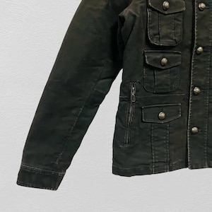 May include: Dark olive green jacket with multiple front pockets and decorative buttons. The jacket features a zipper pocket and buttoned flap pockets. The sleeves are long and the jacket appears to be made of a durable material.