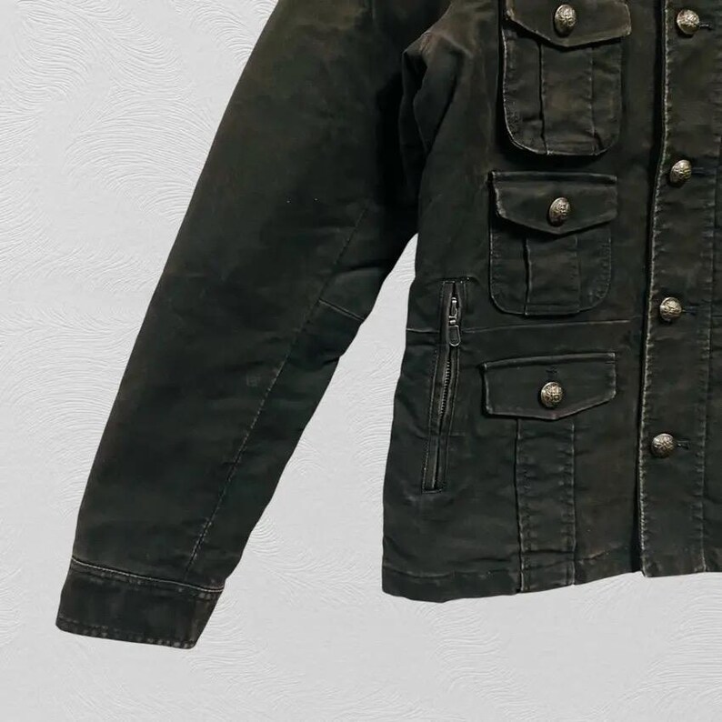 May include: Dark olive green jacket with multiple front pockets and decorative buttons. The jacket features a zipper pocket and buttoned flap pockets. The sleeves are long and the jacket appears to be made of a durable material.