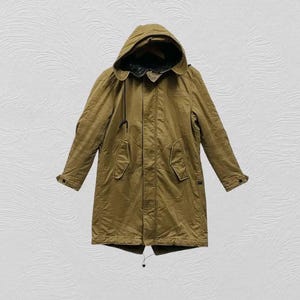 May include: A long, olive-tan parka jacket featuring a large hood with a dark interior lining, a front zipper closure, two slanted flap pockets on the chest, and long sleeves with buttoned cuffs. The coat has a structured, utilitarian design.