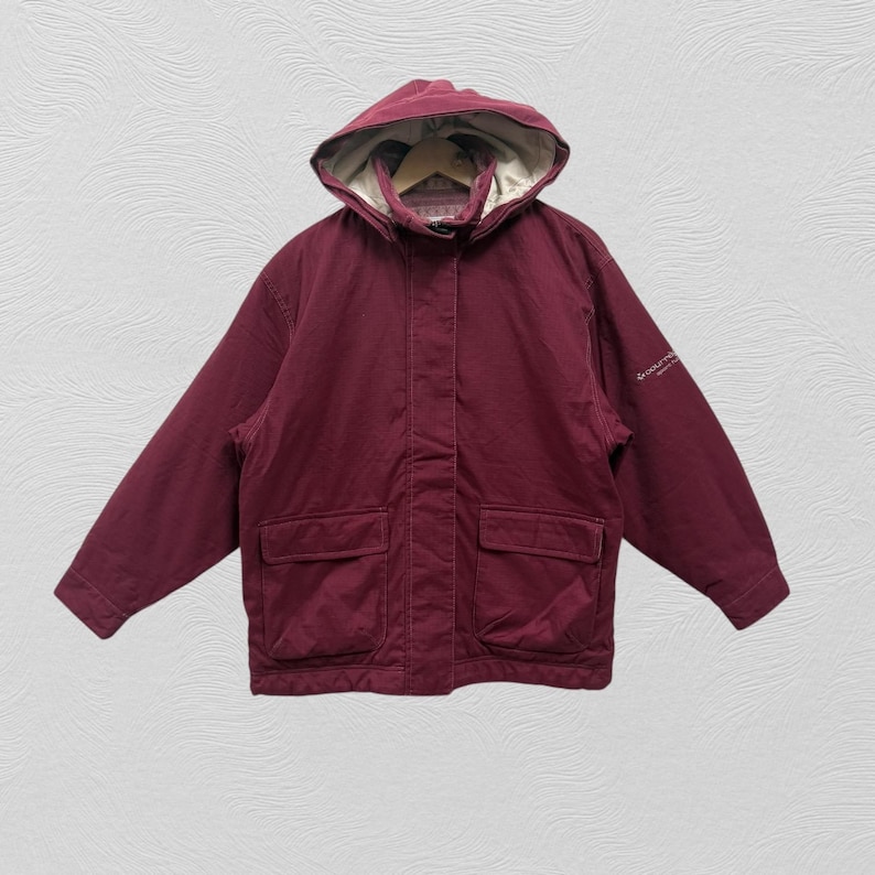 May include: A maroon hooded jacket with two front pockets. The jacket has a light-colored lining and a drawstring hood. The sleeves are long, and the jacket appears to be made of a durable fabric. The jacket is displayed against a textured background.