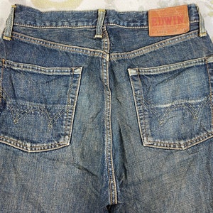 May include: Close-up of the back of blue denim jeans. The jeans have two back pockets with decorative stitching, a brown leather patch with the brand name "EDWIN", and an orange tag. The denim has a worn, faded appearance.