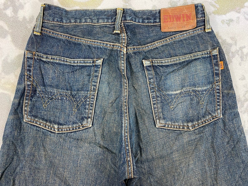 May include: Close-up of the back of blue denim jeans. The jeans have two back pockets with decorative stitching, a brown leather patch with the brand name "EDWIN", and an orange tag. The denim has a worn, faded appearance.