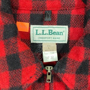 Op de afbeelding: Close-up van een rood en zwart geruit jack met ritssluiting. Het jack heeft een label met "L.L.Bean, Freeport, Maine". Een klein oranje label met het nummer "J536" is bevestigd. De jas is gemarkeerd met "L" en "Made in USA".