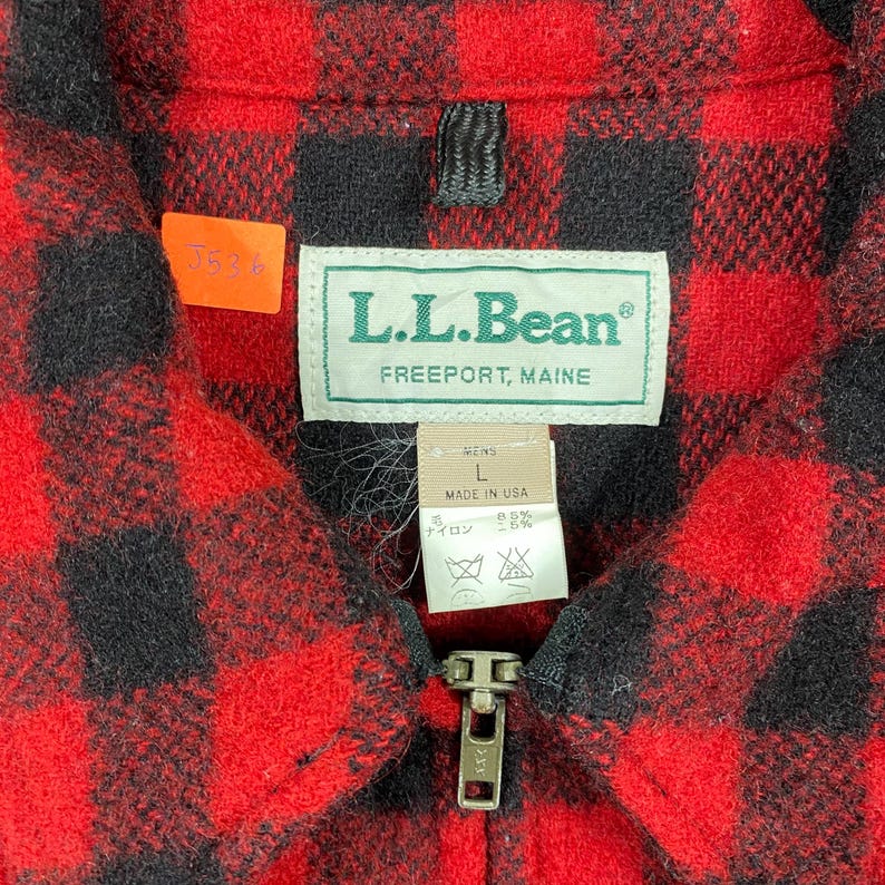 Op de afbeelding: Close-up van een rood en zwart geruit jack met ritssluiting. Het jack heeft een label met "L.L.Bean, Freeport, Maine". Een klein oranje label met het nummer "J536" is bevestigd. De jas is gemarkeerd met "L" en "Made in USA".