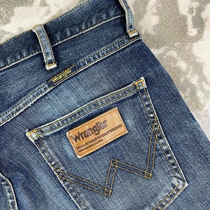 May include: A pair of blue denim jeans with a brown leather patch on the back pocket that says "Wrangler. Wrangler Famous For Quality Workwear. Greensboro, N.C. U.S.A."