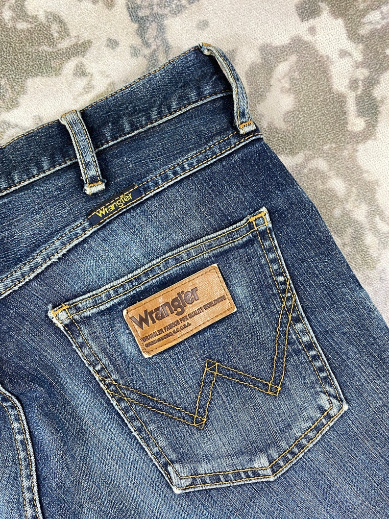 May include: A pair of blue denim jeans with a brown leather patch on the back pocket that says "Wrangler. Wrangler Famous For Quality Workwear. Greensboro, N.C. U.S.A."