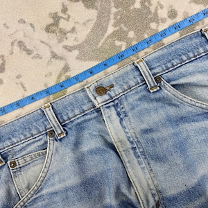 K&ouml;nnte beinhalten: Hellblaue Jeans mit Knopfverschluss und Taschen. Ein blaues Ma&szlig;band liegt &uuml;ber der Jeans und zeigt eine Messung von etwa 43 cm. Die Jeans hat eine verwaschene Optik.