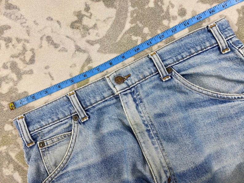 K&ouml;nnte beinhalten: Hellblaue Jeans mit Knopfverschluss und Taschen. Ein blaues Ma&szlig;band liegt &uuml;ber der Jeans und zeigt eine Messung von etwa 43 cm. Die Jeans hat eine verwaschene Optik.