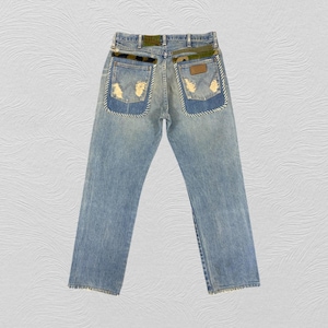Peut inclure: Un jean en denim bleu clair avec un aspect délavé. Les poches arrière sont recouvertes de tissu camouflage et présentent un détail de couture blanche sur les bords.