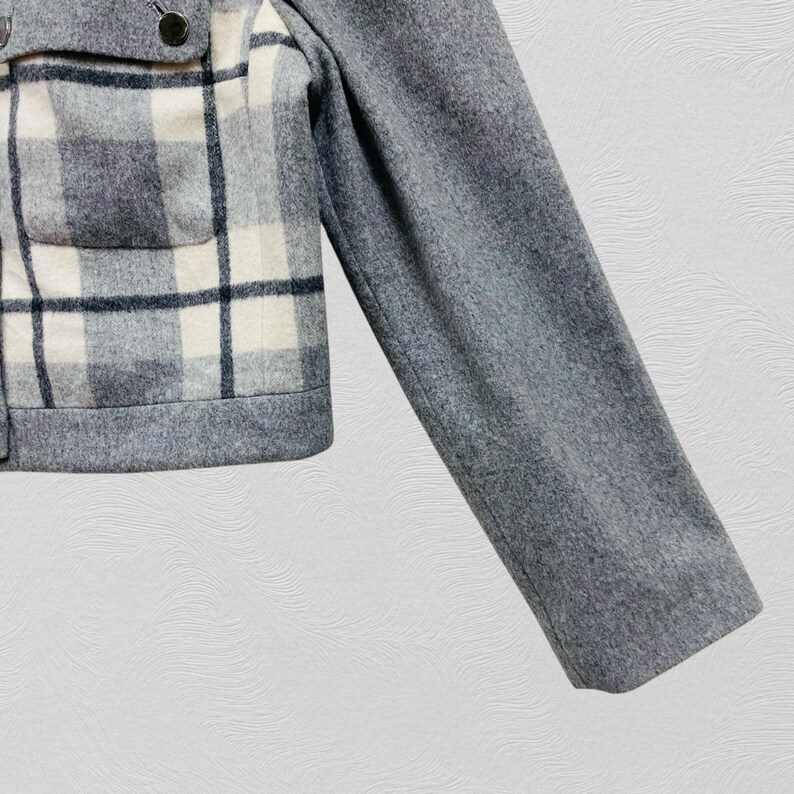 K&ouml;nnte beinhalten: Graue Wolljacke mit einem Karomuster in Wei&szlig;, Grau und Schwarz. Die Jacke hat einen Knopfverschluss und eine einzelne Tasche. Der &Auml;rmel ist einfarbig grau.