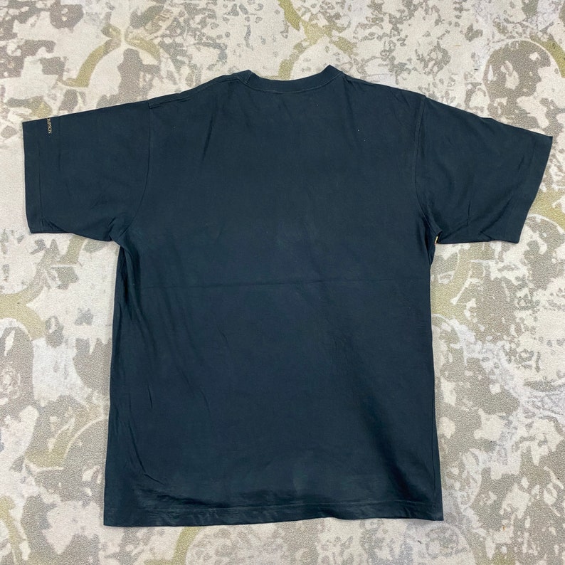 Pode incluir: Uma camiseta preta com gola redonda e mangas curtas. A camisa &eacute; feita de um material macio e tem um design simples e cl&aacute;ssico. A camisa est&aacute; estendida em uma superf&iacute;cie estampada.