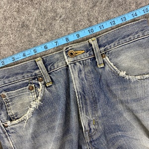 May include: Close-up of light blue denim jeans with distressed pocket edges. A blue measuring tape shows a length of approximately 17 inches. The jeans have a button closure and classic five-pocket design.