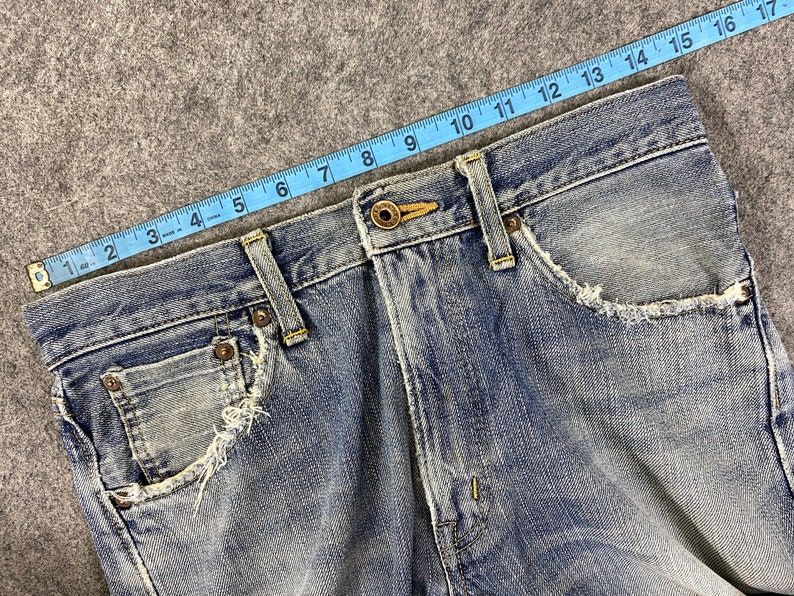 May include: Close-up of light blue denim jeans with distressed pocket edges. A blue measuring tape shows a length of approximately 17 inches. The jeans have a button closure and classic five-pocket design.