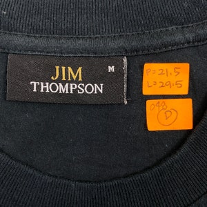 Pode incluir: Close-up de uma gola de camiseta preta com uma etiqueta que diz "JIM THOMPSON" em letras douradas e brancas. A etiqueta tamb&eacute;m tem a letra "M". Duas etiquetas laranja est&atilde;o anexadas com medidas e um "D" circulado.