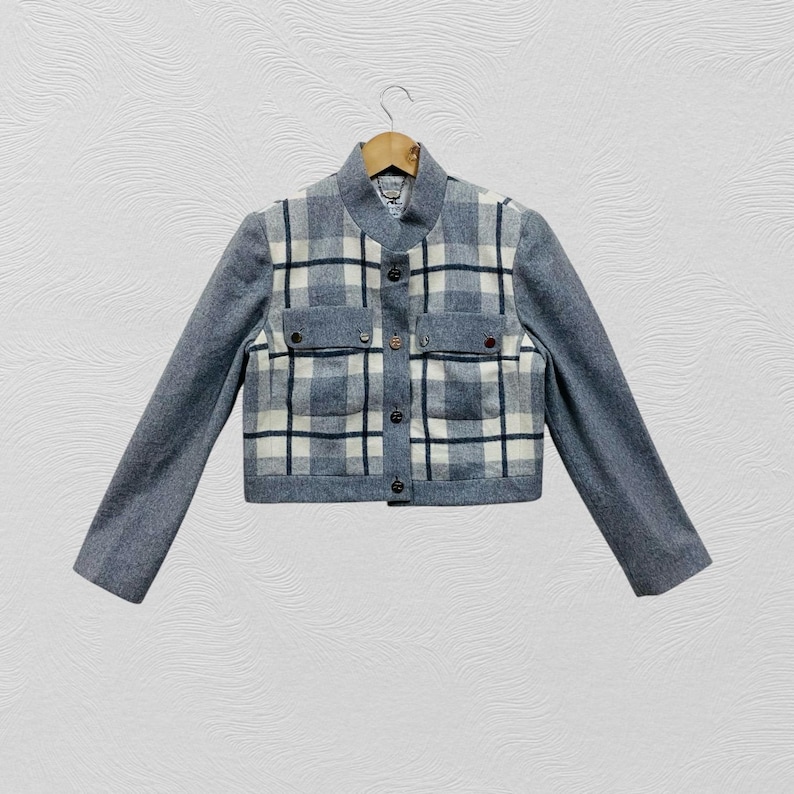 K&ouml;nnte beinhalten: Eine kurze Jacke mit grauem K&ouml;rper und &Auml;rmeln. Die Vorderseite hat ein Karomuster in Wei&szlig;, Marineblau und Grau, mit zwei kleinen Taschen und schwarzen Kn&ouml;pfen. Die Jacke h&auml;ngt an einem Holzb&uuml;gel.