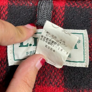 Op de afbeelding: Close-up van een rood-zwarte geruite stof met een label. Het label is wit met groene rand en Japanse tekst. Het label leest "L.L. Bean" en heeft extra Japanse tekst. De stof is een klassiek ruitpatroon.