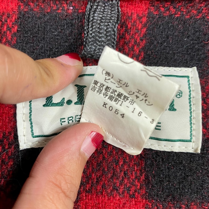 Op de afbeelding: Close-up van een rood-zwarte geruite stof met een label. Het label is wit met groene rand en Japanse tekst. Het label leest "L.L. Bean" en heeft extra Japanse tekst. De stof is een klassiek ruitpatroon.