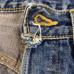 May include: Close-up of a section of blue denim jeans, showcasing the buttonhole and button area. The buttonhole is reinforced with yellow stitching, and the denim has a worn, distressed appearance. Frayed threads and a textured surface are visible.