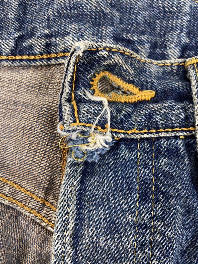 May include: Close-up of a section of blue denim jeans, showcasing the buttonhole and button area. The buttonhole is reinforced with yellow stitching, and the denim has a worn, distressed appearance. Frayed threads and a textured surface are visible.