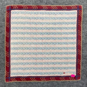 May include: Square handkerchief with a cream-colored center featuring a pattern of blue chevrons. The border is a deep red with a repeating arrow design and a tan outer edge. The brand name "Issey Miyake" is visible.