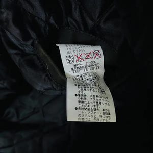 May include: A black garment tag with Japanese text and care symbols. The tag includes washing instructions, a hand wash symbol, and the text "MADE IN CHINA". The tag is attached to a black fabric.