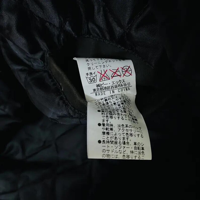 May include: A black garment tag with Japanese text and care symbols. The tag includes washing instructions, a hand wash symbol, and the text "MADE IN CHINA". The tag is attached to a black fabric.
