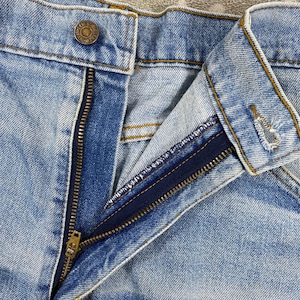 K&ouml;nnte beinhalten: Nahaufnahme einer hellblauen Jeans mit einem teilweise ge&ouml;ffneten Messingrei&szlig;verschluss. Die Jeans hat einen Knopfverschluss am Bund und sichtbare N&auml;hte. Der Stoff zeigt einen verblichenen Wascheffekt.