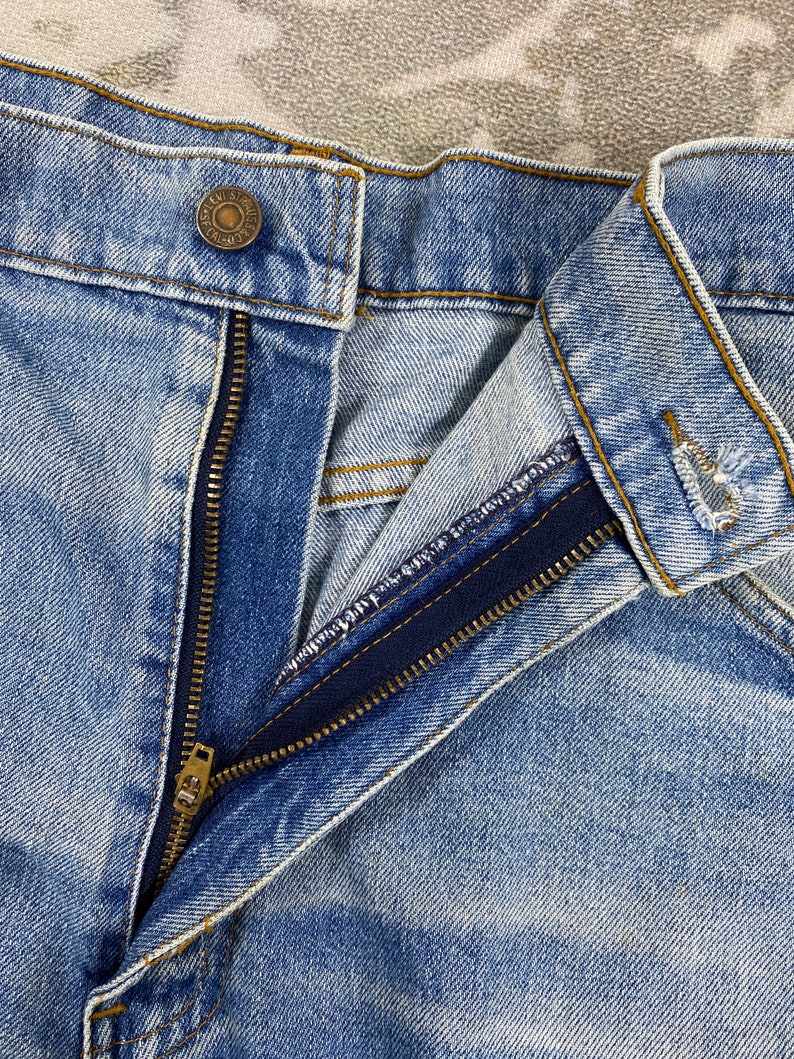 K&ouml;nnte beinhalten: Nahaufnahme einer hellblauen Jeans mit einem teilweise ge&ouml;ffneten Messingrei&szlig;verschluss. Die Jeans hat einen Knopfverschluss am Bund und sichtbare N&auml;hte. Der Stoff zeigt einen verblichenen Wascheffekt.