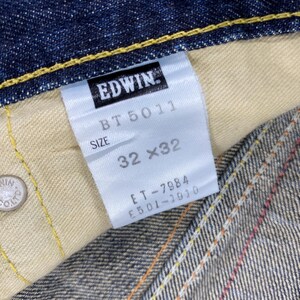 May include: Dark blue denim fabric with yellow contrast stitching and a silver metal rivet embossed with Edwin Co. Ltd. A white interior tag displays the brand name Edwin, model BT 5011, size 32x32, and additional serial numbers ET-7984 and E501-1910.
