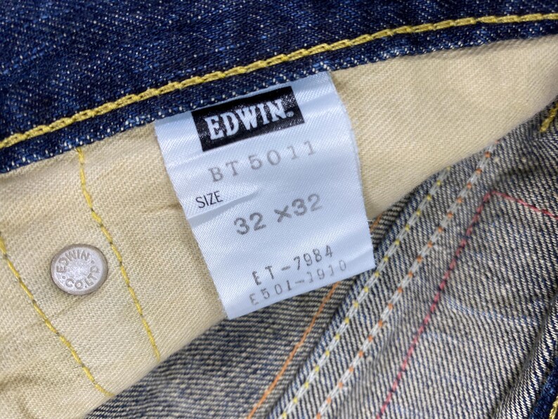May include: Dark blue denim fabric with yellow contrast stitching and a silver metal rivet embossed with Edwin Co. Ltd. A white interior tag displays the brand name Edwin, model BT 5011, size 32x32, and additional serial numbers ET-7984 and E501-1910.