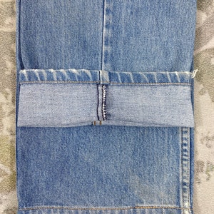 K&ouml;nnte beinhalten: Nahaufnahme einer hellblauen Jeans mit umgeschlagenem Saum. Die Jeans hat ein klassisches F&uuml;nf-Taschen-Design und einen leicht ausgefransten Saum. Der Stoff weist subtile Ausbleichungen und Abnutzungserscheinungen auf, was auf einen Vintage- oder beliebten Stil hindeutet.